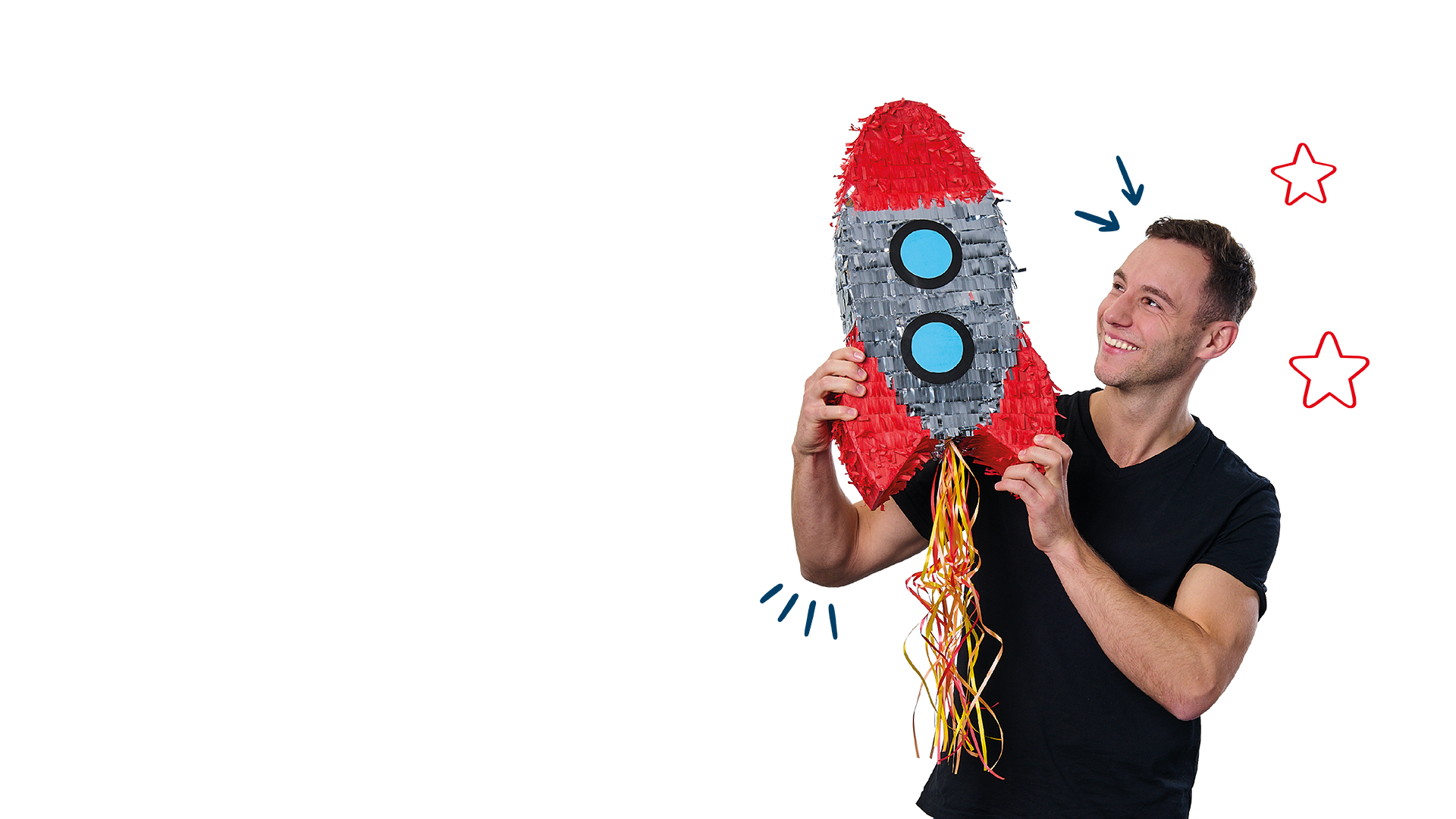 Lächelnder junger Mann mit kurzen, dunklen Haaren trägt ein schwarzes T-Shirt und hält eine bunte Raketen-Piñata in den Händen. Er betrachtet sie fröhlich, als ob er sie gleich starten lassen würde.